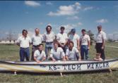 Color photograph of eleven individuals standing and kneeling behind a banner containing the text "The KS-Tech Merger"