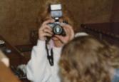 Color photograph of two individuals. One individual is looking through a camera, and the other has their head bent down out of frame.