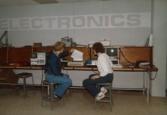 Color photograph of two individuals sitting at a desk, examining a sheet of paper being printed out.