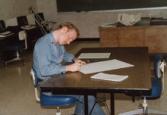 Color photograph of an individual sitting at a desk and writing on a sheet of paper.