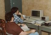 Color photograph of two individuals sitting and working at a computer.
