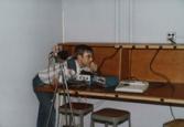 Color photograph of an individual crouching over a desk, examining equipment.